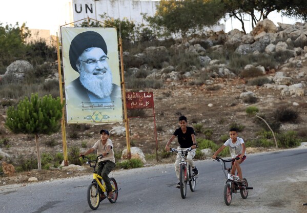 Children ride their bicycles past by a poster of Hezbollah leader Sayyed Hassan Nasrallah at the Lebanese-Israeli border village of Marwaheen, south of Lebanon, Wednesday, July 12, 2023. An explosion near Lebanon's border with Israel slightly wounded at least three members of the militant Hezbollah group, a Lebanese security official said. (AP Photo/Mohammed Zaatari)